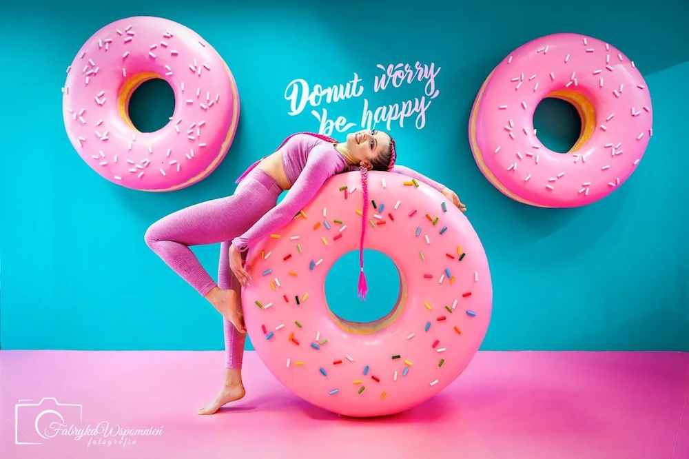 obraz przedstawia jedną z instalacji dostępnych do zobaczenia w Muzeum Szczęścia i Iluzji w Szczecinie- Be Happy. na obrazie jest dziewczyna opierająca się o wielkie różowe pączki z dziurą- donuty