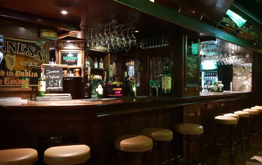 obraz przedstawia fragment wnętrza- ladę, wiszące kieliszki do wina, nalewaki i wysokie krzesła obciągnięte jasną skórą