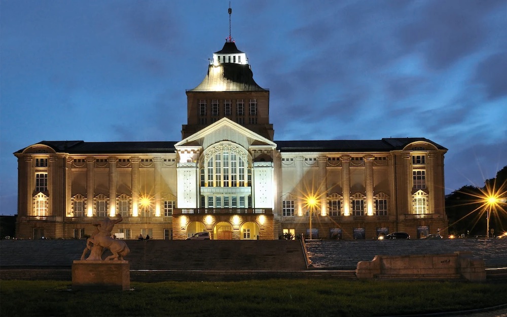 Obraz przedstawia przednią ścianę Muzeum Narodowego w Szczecinie wieczorną porą; na ściany pada ciepłe światło