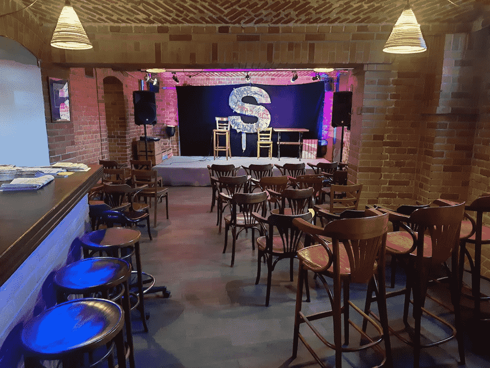 obrazek przedstawia wnętrze szczecińskiej Piwnicy Kany. Na środku widoczna jest niewielka scena oraz większa ilość krzeseł