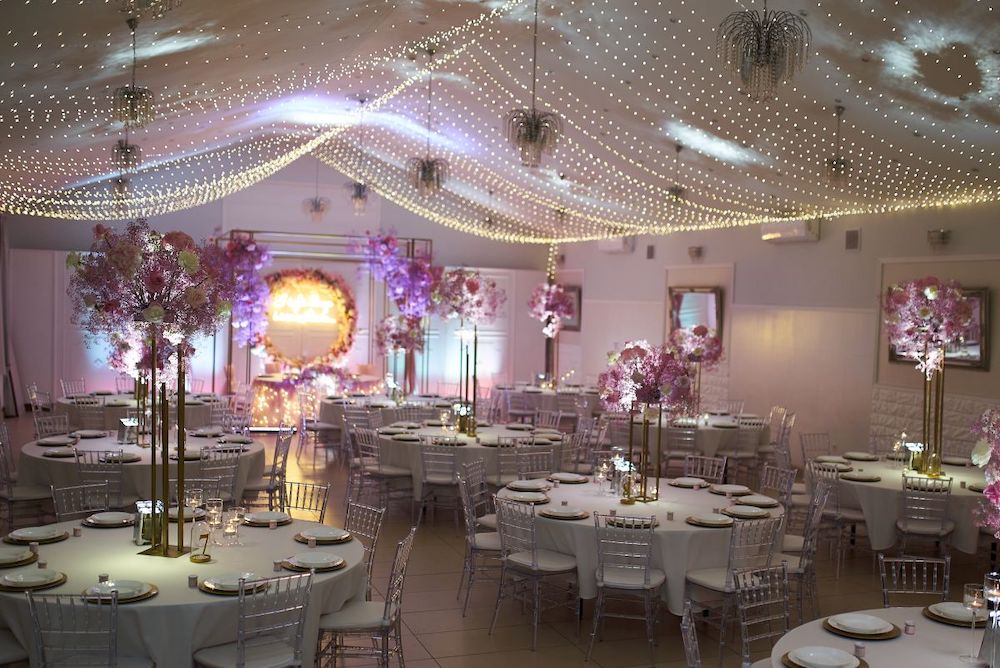 obrazek przedstawia jasno-różowe wnętrze sali Lila Park w Szczecinie; okrągłe stoliki i wysokie różowe instalacje kwiatowe w otoczeniu wiszących fal lampeczek;